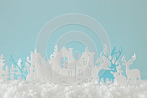 Magical Christmas paper cut winter background landscape with houses, trees, deer and snow in front of pastel blue background.
