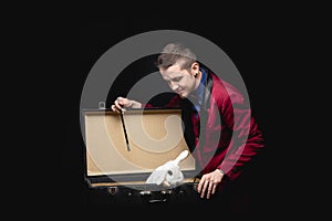 Magician shows trick with disappearance white rabbit in suitcase magic wand, black background