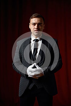 Magician wearing suit standing with cane on stage decorated red velvet curtain