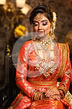 Magnificent young Indian bride in luxurious bridal costume with makeup and heavy jewellery is sitting in a chair