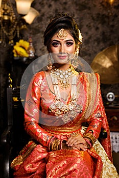 Magnificent young Indian bride in luxurious bridal costume with makeup and heavy jewellery is sitting in a chair in with classic