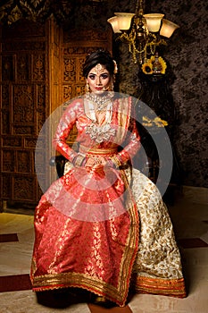 Magnificent young Indian bride in luxurious dress and precious jewellery is sitting in a chair in a luxury apartment. Classic