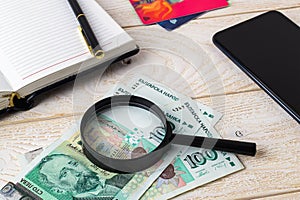 Magnifier on bulgarian levs near pen, note book, credit cards and smartphone on a white wooden surface. Check the authenticity of