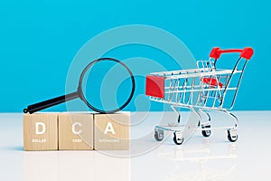 Magnifying glass and DCA word on a wooden cube in idea Dollar Cost Averaging investment strategy near empty shopping cart, Saving