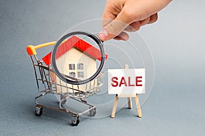 Magnifying glass is looking at the Supermarket cart with houses and a Sale Poster. The concept of selling a home, real estate