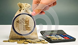 Magnifying glass looks at money bag with coins and a calculator. Profit calculation and income analysis. Interest rates. photo