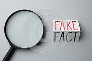 Magnifying glass near sheet of paper with words Fact and Fake on light grey background, flat lay