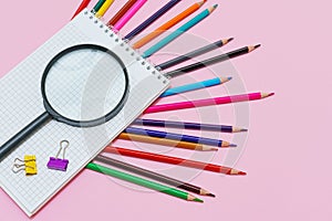 Magnifying glass, pencils and a blank white notepad on a spring on a pink