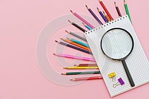 Magnifying glass, pencils and a blank white notepad on a spring on a pink