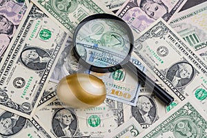 Magnifying glass with shiny golden egg under pile of US America dollar banknotes money metaphor of precious wealth, finding good