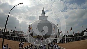 main entrance to the Buddhist sanctuary Ruwanwelisaya stupa, a crowd of people on the backlit faceless in white and flags in the w