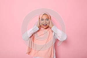 Happy religious Muslim woman in pink hijab holding her head with hands and smiling with toothy smile looking at camera with