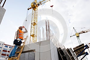 Maintenance worker man with safety helmet and orange vest climbing wood step ladder and holding toolbox at construction site