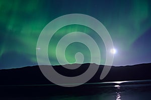 Majestic aurora borealis dancing beside full moon over mountain and calm fjord