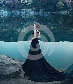 A majestic lady, a dark queen, stands on the background of a river and rocks, in a long black dress. The brunette girl