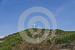 A majestic wooden cross rises over a hill overgrown with coniferous and deciduous trees against the blue sky near Blagoevgrad