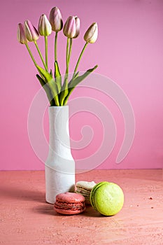 Makaroons on a pink background next to a bouquet of tulips in a white vase and copy space