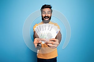 Make dream come true. Portrait happy indian man showing new car key and money cash dollars in hand, blue background