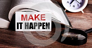 MAKE IT HAPPEN written on a white card near an open book, an alarm clock and a magnifying glass on a wooden table. Call to action