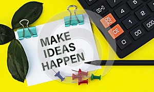 MAKE IDEAS HAPPEN - words on a white sheet with clips on a yellow background with a calculator, buttons and pencil