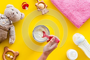 Make mixture for feed baby. Spoon with food in hand near toys and accessories on yellow background top view pattern