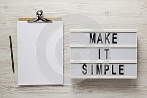 `Make it simple` words on a lightbox, clipboard with blank sheet of paper on a white wooden background, overhead view. Top view,