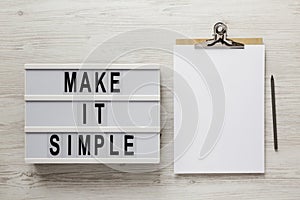 `Make it simple` words on a lightbox, clipboard with blank sheet of paper on a white wooden background, view from above. Top vie