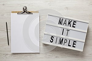 `Make it simple` words on a lightbox, clipboard with blank sheet of paper on a white wooden surface, overhead view. Top view, fr