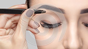 Make-up artist plucks eyebrows with tweezers to a woman with curly brown hair and nude make-up. Beautiful thick eyebrows close up