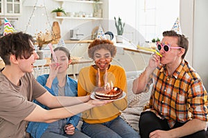 Make a wish. Woman wearing party cap blowing out burning candles on birthday cake. Happy Birthday party. Group of