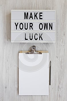 `Make your own luck` on a lightbox, clipboard with blank sheet of paper on a white wooden background, top view. Flat lay, from