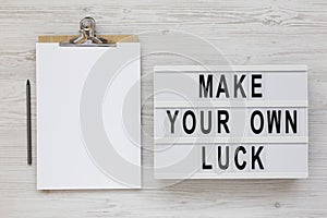 `Make your own luck` on a lightbox, clipboard with blank sheet of paper on a white wooden surface, top view. Flat lay, from abov