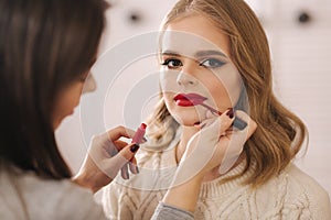 Makeup artist applies red lipstick. Hand of make-up master, painting lips of young beauty blond hair model. Make up in