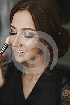 Makeup artist applying makeup on face of a beautiful and cheerful brunette model girl with charming smile and closed