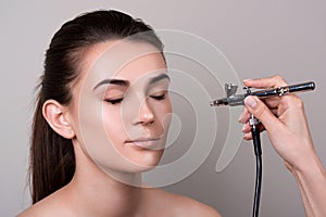 Makeup artist using airbrush. Fashion brunette model with hair tail over gray background. Professional makeup with aerograph. Girl photo