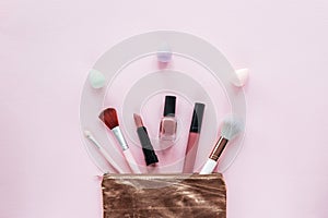 Makeup brushes, nail polish, lipstick and colorful sponges in golden cosmetic bag on pink background. Top view, flat lay