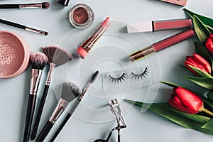 Makeup cosmetics and red peony flowers on a blue background. Brushes, shadow palette, lipstick and eyelashes for the