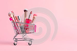 Makeup products in shopping cart on pink background. Lipstick, mascara, lip gloss, brushes, nail polish. Shopping cart full of