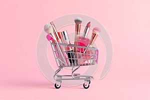 Makeup products in shopping cart on pink background. Lipstick, mascara, lip gloss, brushes, nail polish. Shopping cart full of