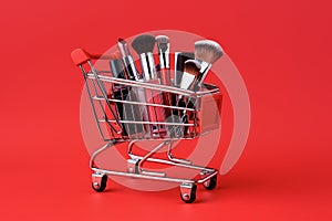 Makeup products in shopping cart on red background. Lipstick, mascara, lip gloss, brushes, nail polish. Shopping cart full of