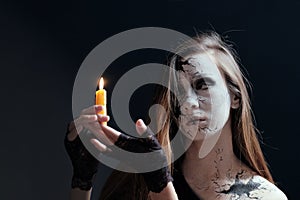 Makeup in the style of Halloween. A young girl with long hair with cracks painted on her face is holding a burning candle. Dark ba