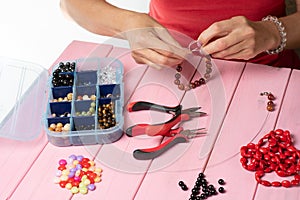 Making a bracelet of colorful beads. Female hands with a tool on a pink wooden table.