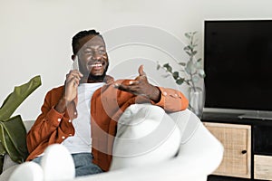Making call. Happy african american man talking on smartphone and gesturing, resting on sofa at home, free space