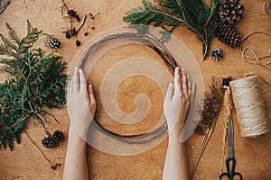 Making christmas wreath. Hands holding wooden circle and fir branches, pine cones, thread, scissors, herbs on wooden table.