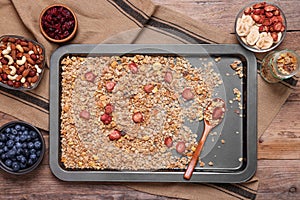 Making granola. Baking tray with mixture of oat flakes, other ingredients and spoon on wooden table, flat lay