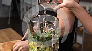 Making green juice at home: Female hands pouring water into a blender in the kitchen. Unrecognizable fitness woman in