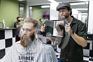 Making haircut look perfect. Young bearded man getting haircut by hairdresser while sitting in chair at barbershop