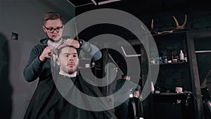 Making haircut look perfect. Young bearded man getting haircut by hairdresser while sitting in chair at barbershop