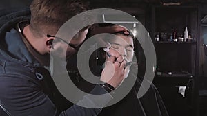 Making haircut look perfect. Young bearded man getting haircut by hairdresser while sitting in chair at barbershop