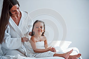 Making hairstyle by using comb. Young mother with her daugher have beauty day indoors in white room